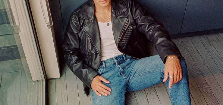  Man sitting on the floor wearing a black leather jacket, white tank top, and blue jeans, accessorized with rings and a necklace.