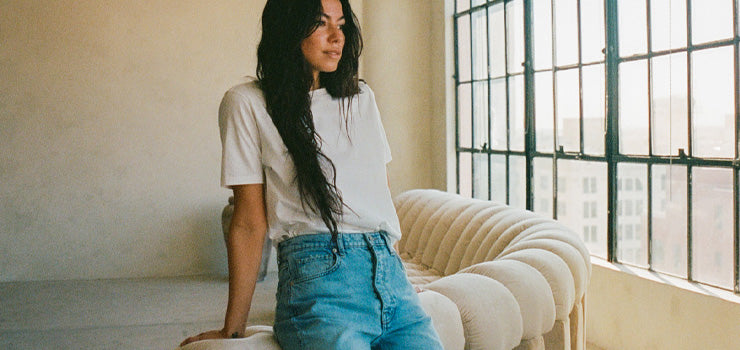 Woman in a white t-shirt and high-waisted blue jeans sitting on a cream tufted sofa in a sunlit loft with large industrial windows.